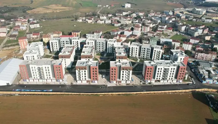 KİPTAŞ konut projesinin havadan görünümü: kırmızı-beyaz cepheli bloklar, arkada mevcut mahalle, önde tarla