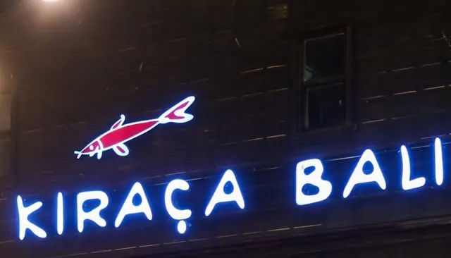 Kıraça Balık'ın gece aydınlatılmış cephesi: ahşap duvarda neon balık figürü ve işletmenin adı